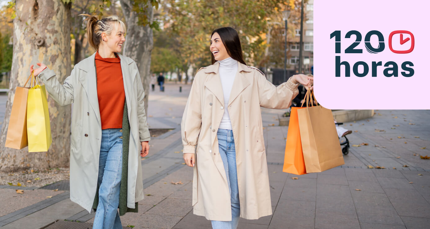 Dos mujeres vestidas con gabardinas, paseando por la calle, sonriendo y sosteniendo bolsas de compras.