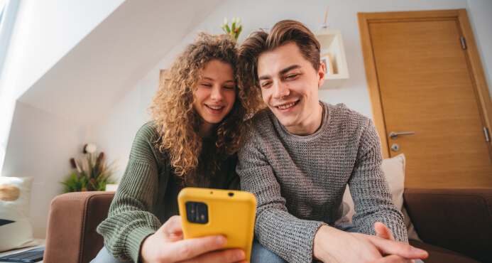 Joven sonriendo mientras usa la app móvil de su banco en un celular con funda amarilla, junto a su hermano.