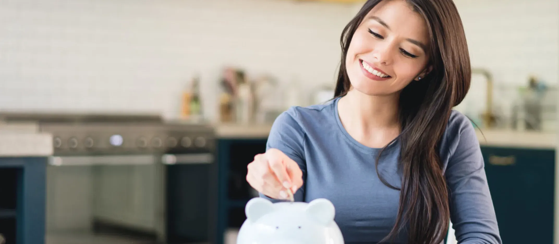 Persona colocando una moneda en una alcancía con forma de cerdito sobre una mesa en una cocina moderna.