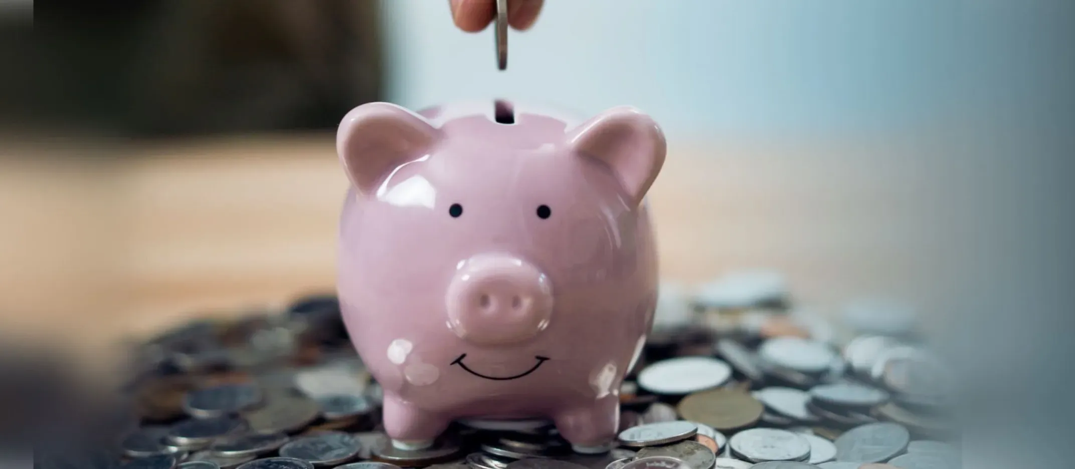 Alcancía en forma de cerdito rosa rodeada de monedas, con una mano depositando una moneda en la ranura, representando ahorro para el retiro y Afore.