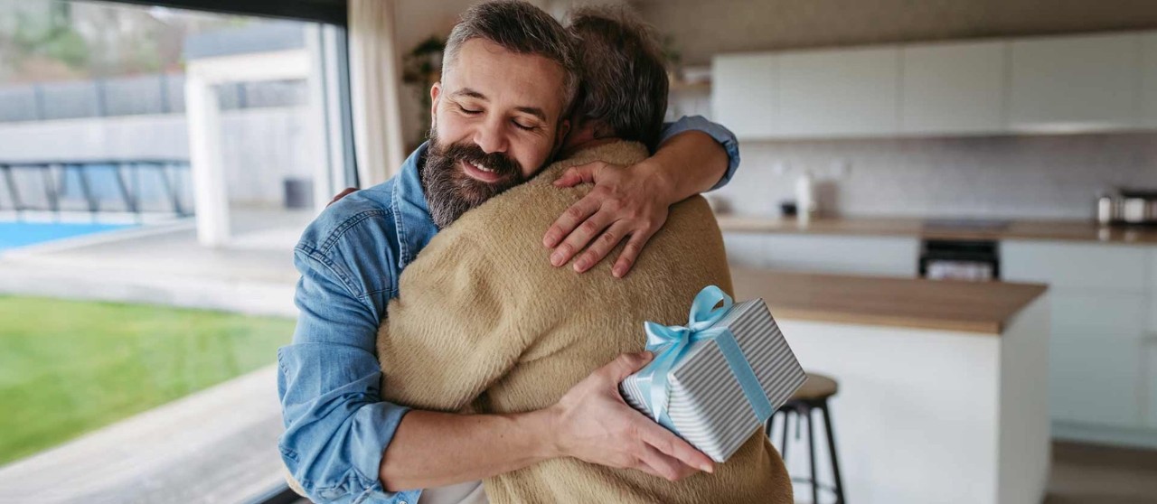 Papá abrazando a su hijo en una cocina moderna mientras sostiene un regalo envuelto con moño azul.