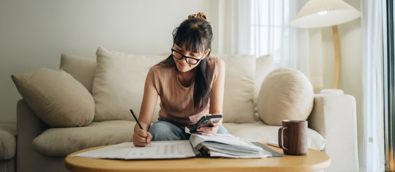 Persona sentada en un sofá escribiendo en un cuaderno mientras sostiene un teléfono móvil.
