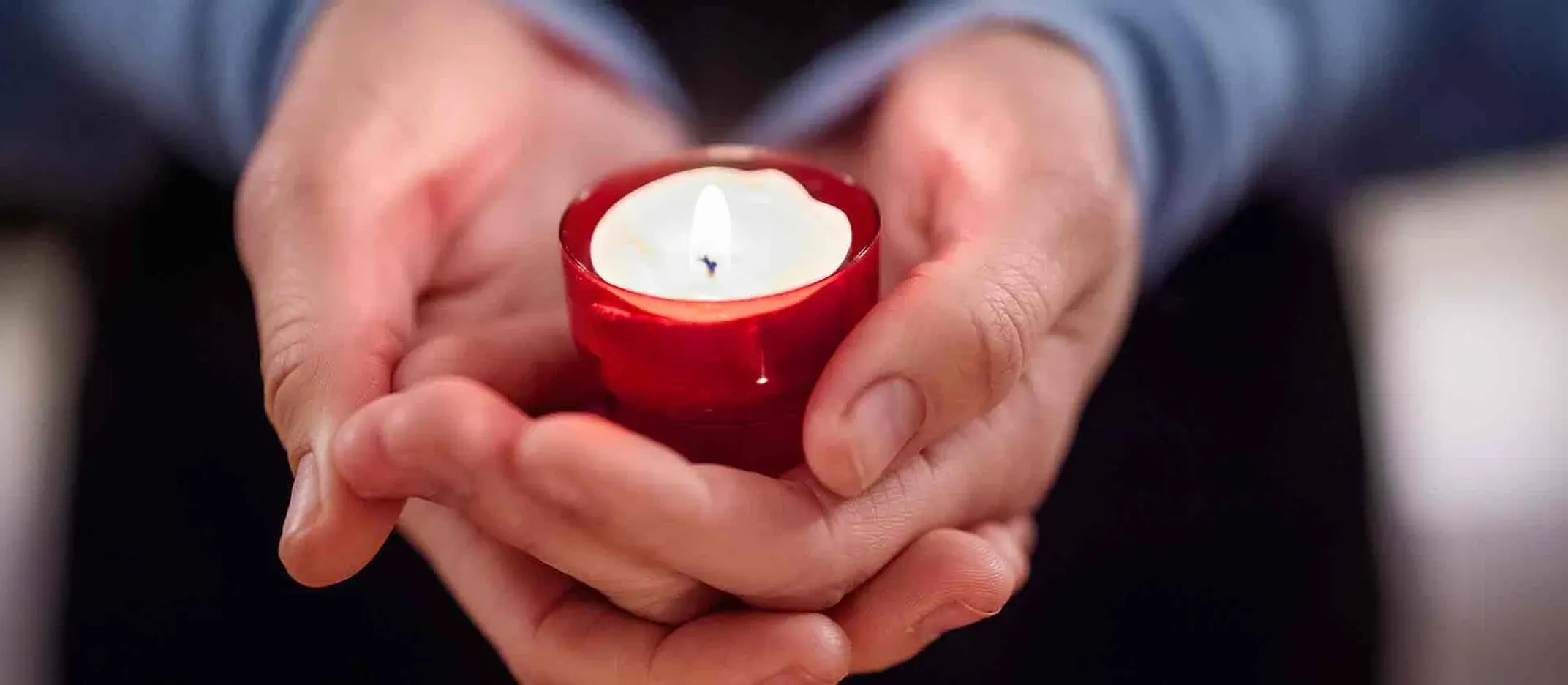 Manos sosteniendo una vela encendida en recipiente rojo, símbolo de recuerdo y homenaje.
