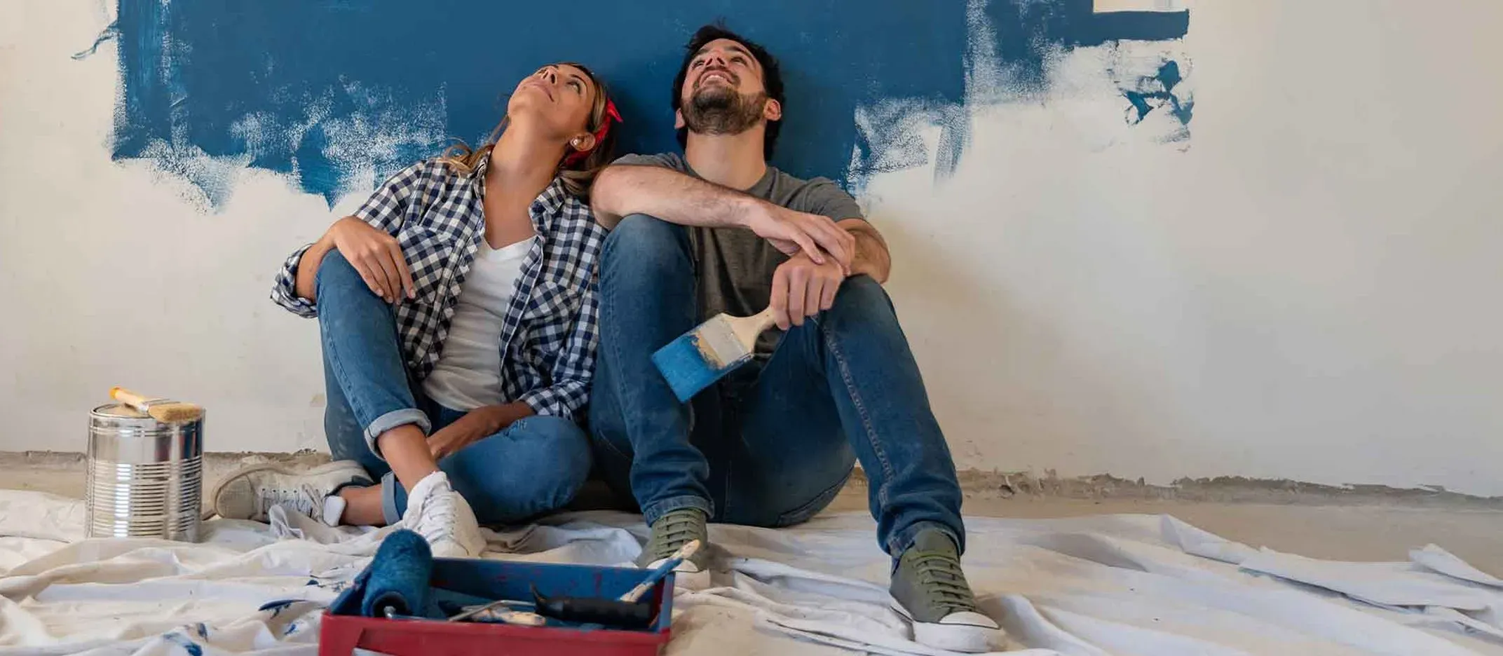 Pareja sentada en el suelo frente a pared parcialmente pintada de azul, con rodillos, brochas y lata de pintura.