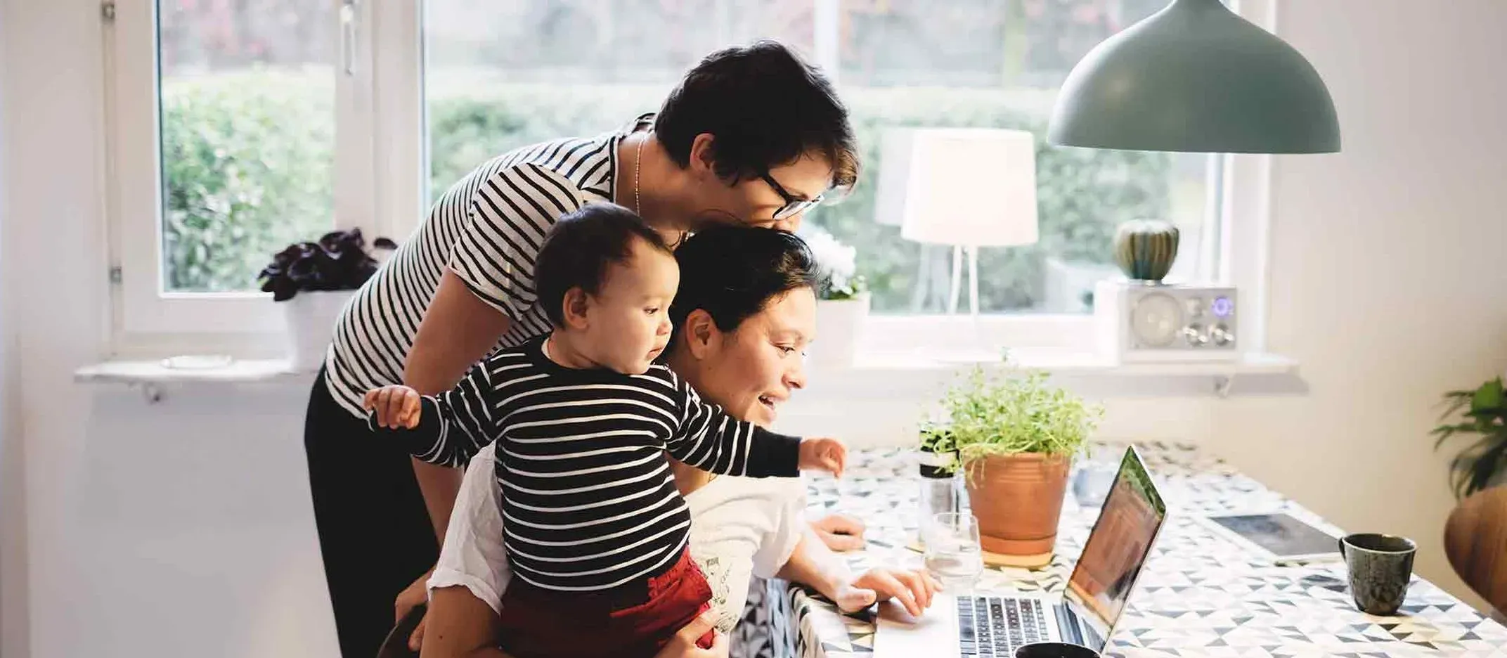 Persona sosteniendo a un bebé mientras revisa una laptop sobre mesa con mantel geométrico, en cocina iluminada con luz natural.