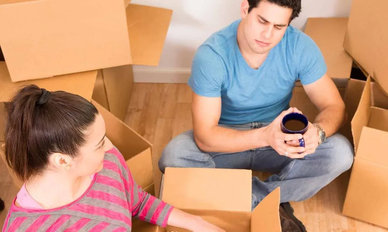Dos personas sentadas en el suelo rodeadas de cajas de cartón abiertas, en proceso de mudanza dentro de una habitación con piso de madera.