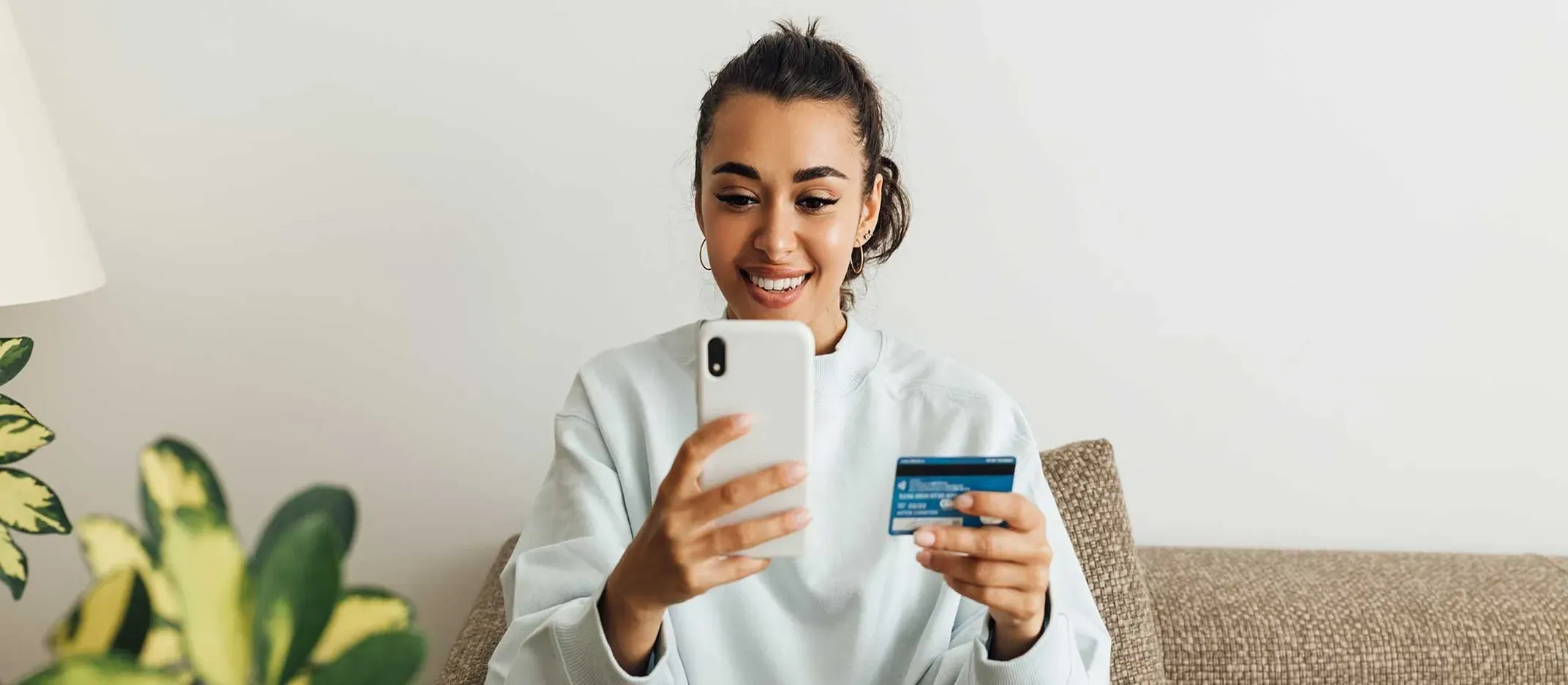 Persona sosteniendo tarjeta bancaria y teléfono móvil, realizando operación financiera en línea desde un sofá con planta decorativa al lado.