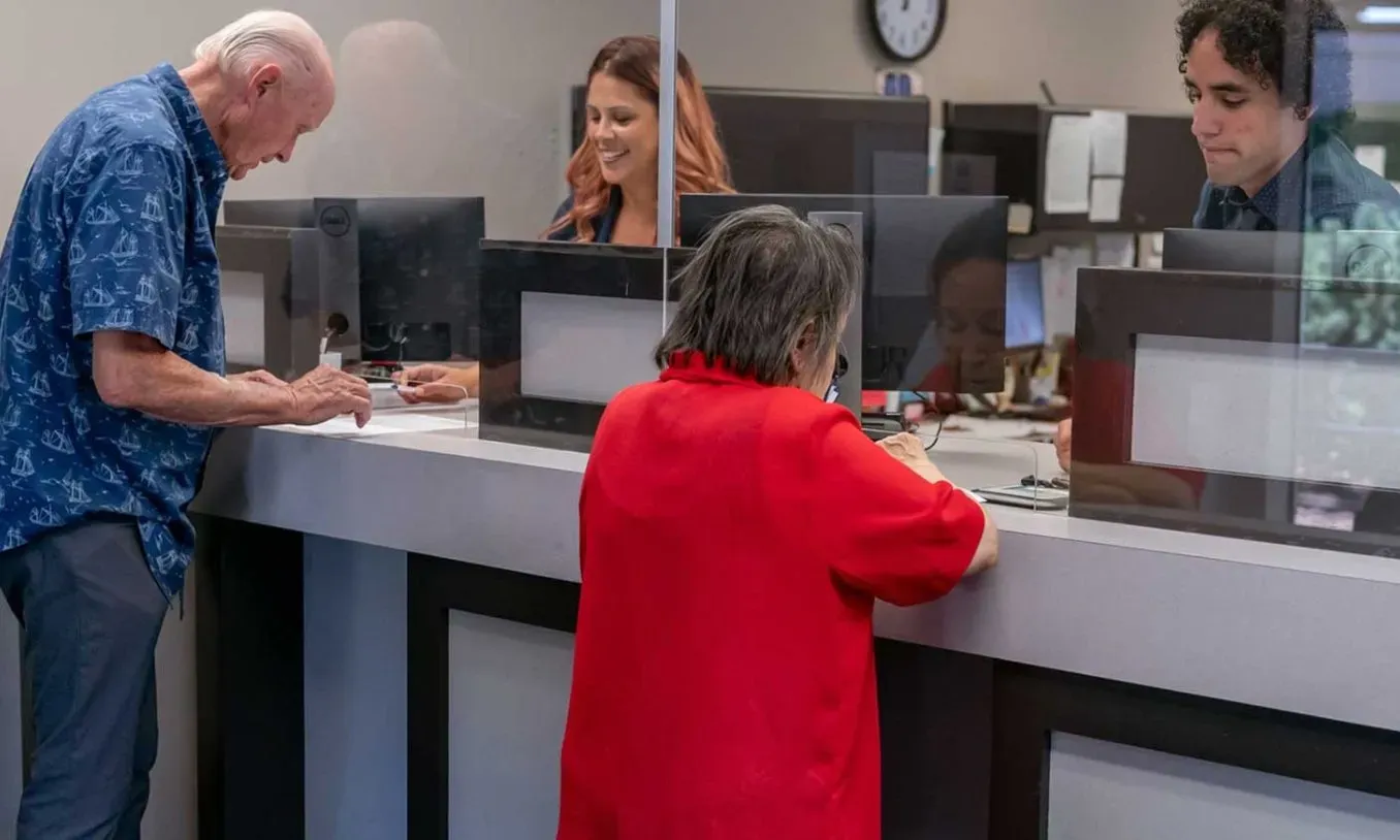 Personas adultas mayores realizando trámites en ventanilla bancaria, con personal atendiendo detrás de mamparas transparentes en una sucursal.