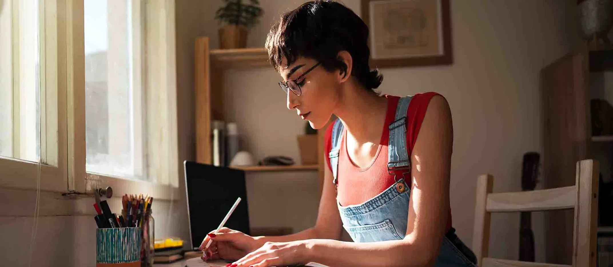 Persona escribiendo en un escritorio junto a una ventana iluminada, con laptop, lápices y estantería de madera en el fondo.