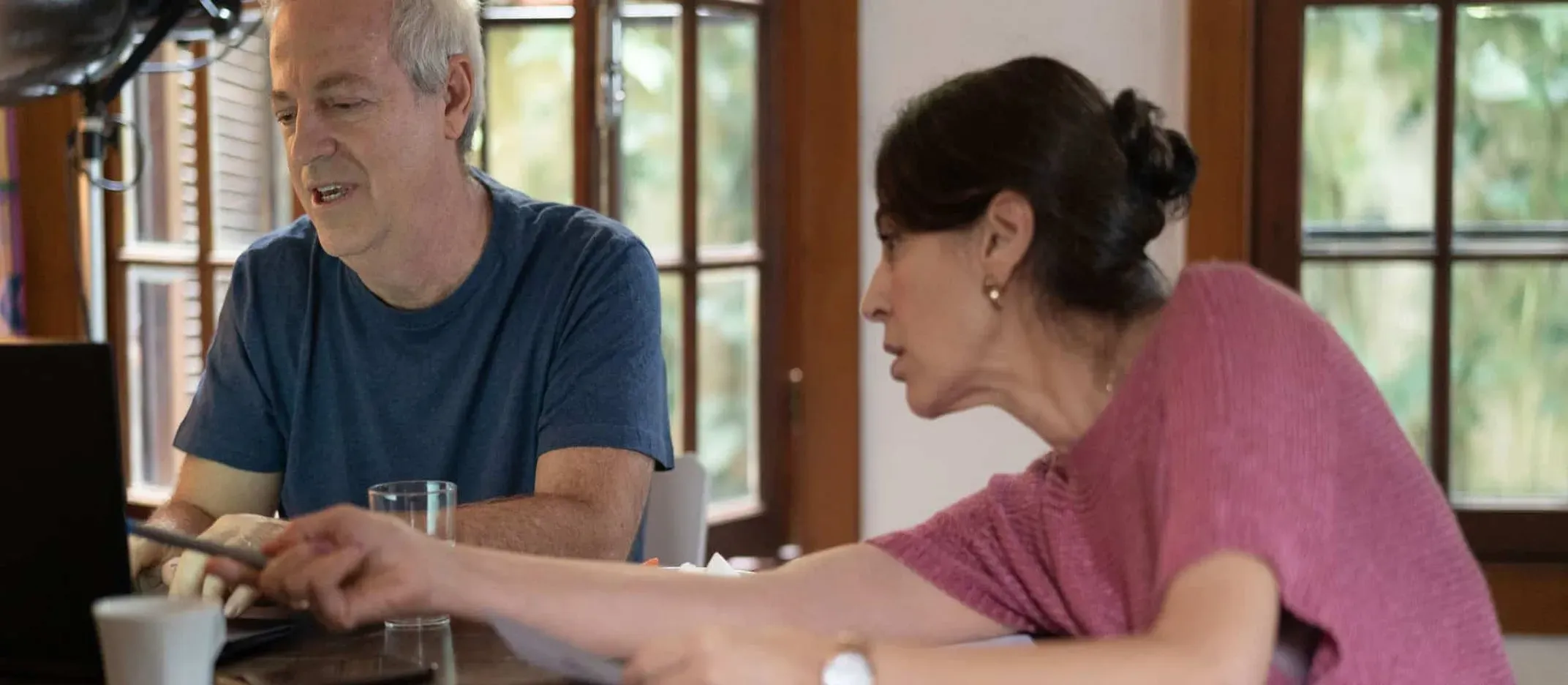 Dos personas revisando documentos y computadora portátil en una mesa de madera, con tazas y vasos, en una habitación iluminada con ventanas grandes.