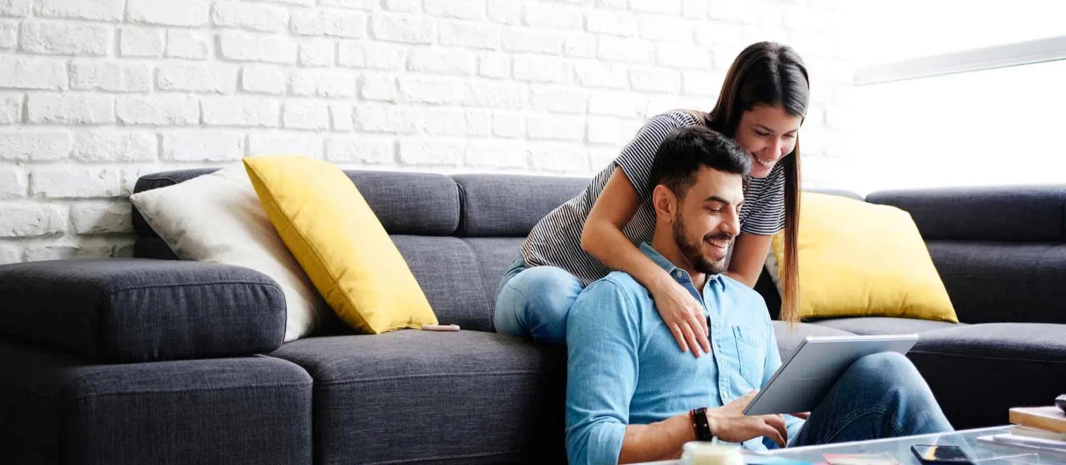 Pareja sentada en sofá gris con cojines amarillos y blancos, revisando una tablet sobre mesa de centro, con pared de ladrillo blanco al fondo.