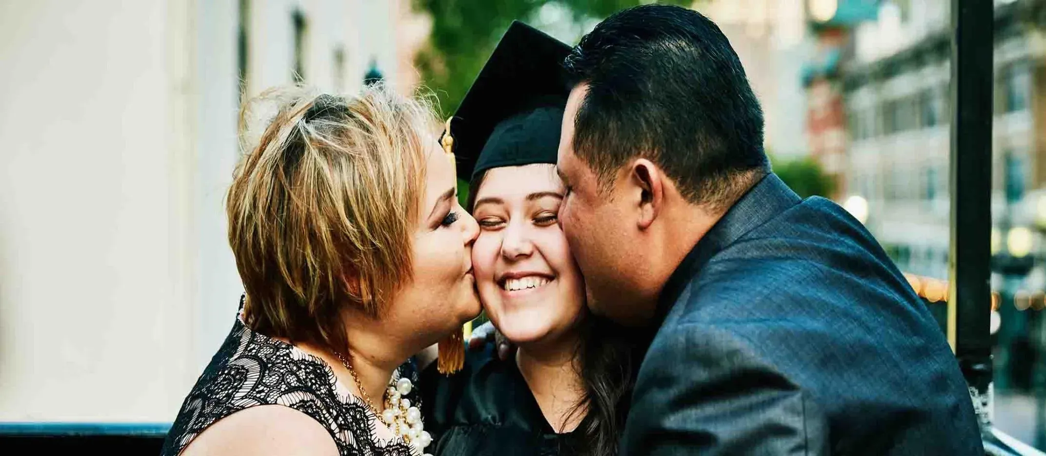 Dos personas abrazando a un graduado con toga y birrete en ceremonia al aire libre, edificios urbanos desenfocados al fondo.