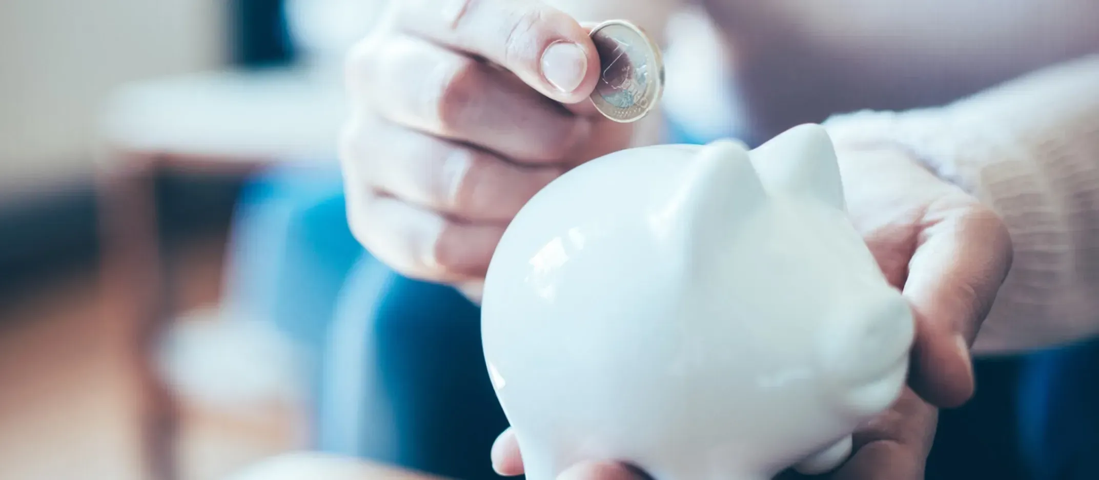 Mano colocando una moneda en una alcancía blanca con forma de cerdito, representando ahorro inicial para comenzar a invertir con poco dinero.