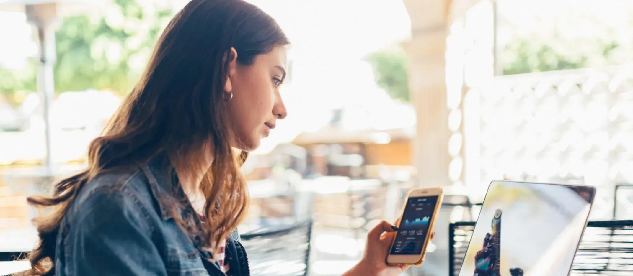 Persona sentada en una mesa al aire libre utilizando un smartphone y una laptop, mostrando gráficos financieros en pantalla, representando análisis de inversiones.