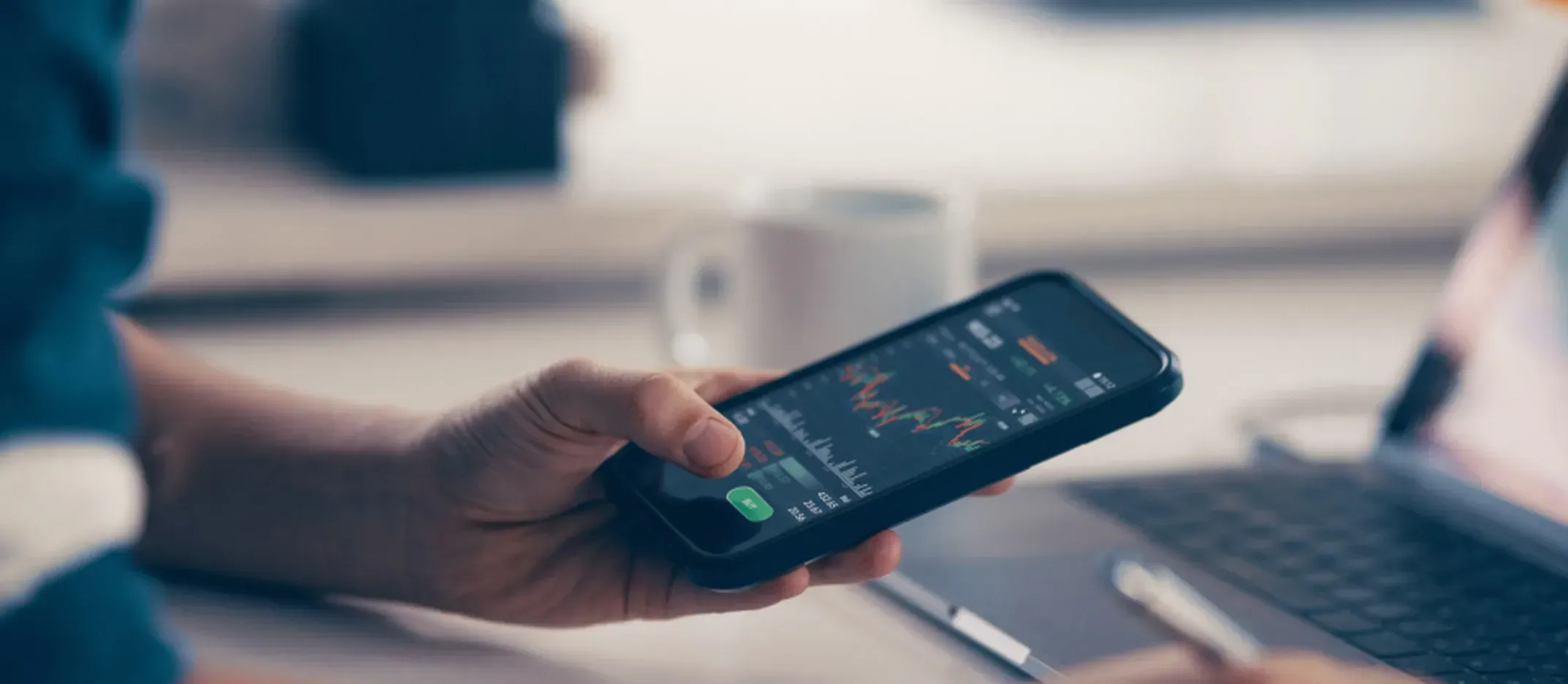 Mano sosteniendo un smartphone con gráficos de mercado y datos bursátiles en pantalla, frente a una computadora portátil en un escritorio, representando análisis financiero e inversiones digitales.
