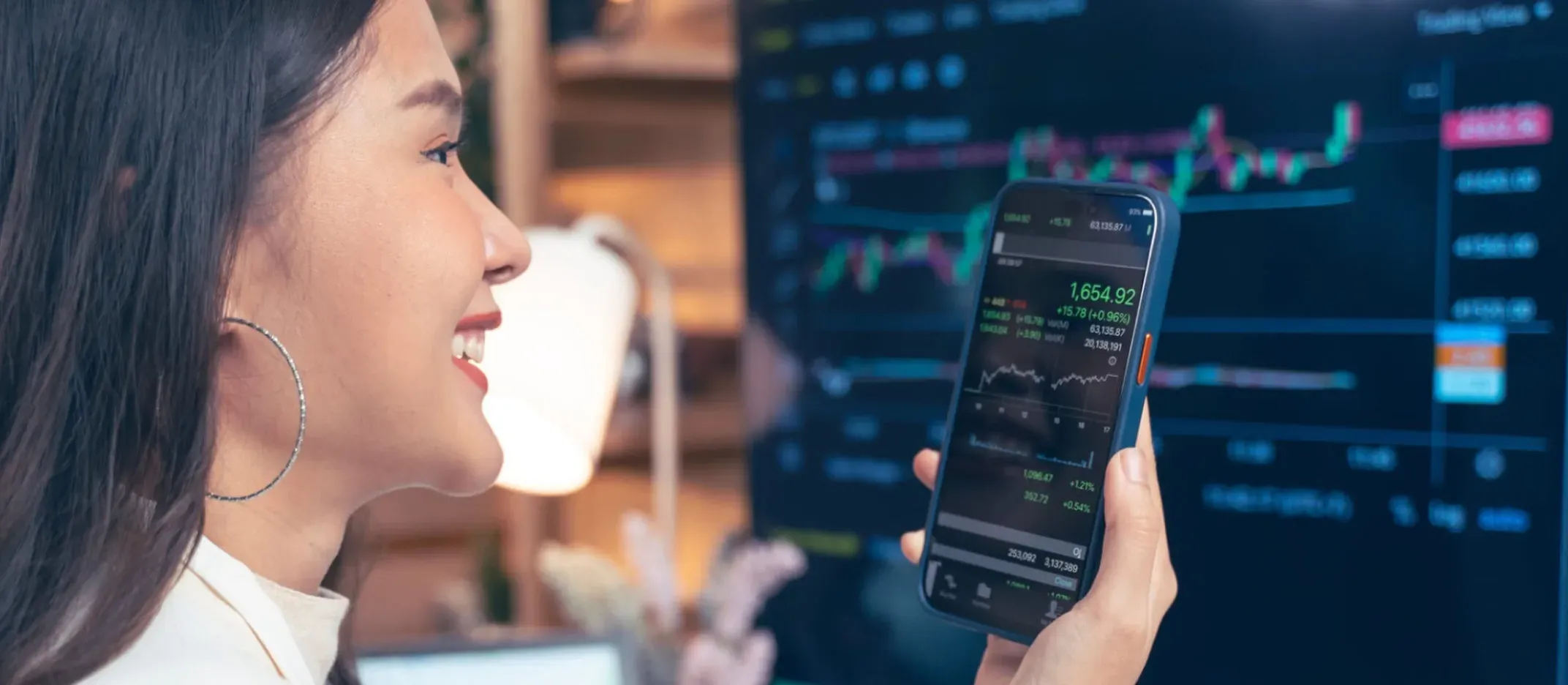 Persona sosteniendo un teléfono móvil con gráficos financieros y datos bursátiles en pantalla, frente a un monitor grande que muestra gráficas de mercado.