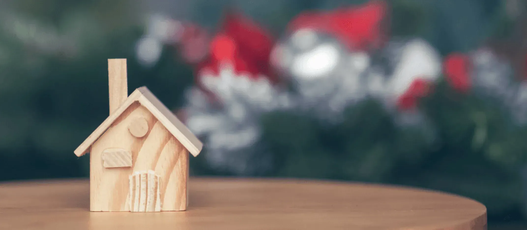 Figura decorativa de madera en forma de casa sobre una mesa redonda, con fondo desenfocado de adornos navideños en tonos rojo, blanco y verde.