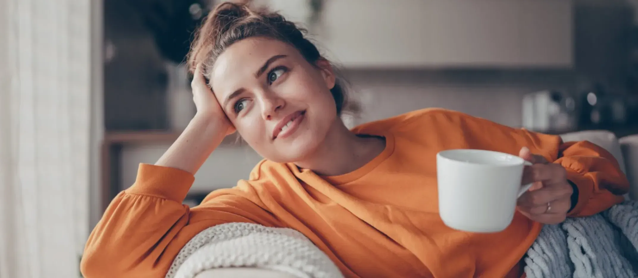 Persona descansando en un sofá con una taza blanca en la mano, usando ropa cómoda y una manta, representando bienestar y hábitos para ahorrar dinero.