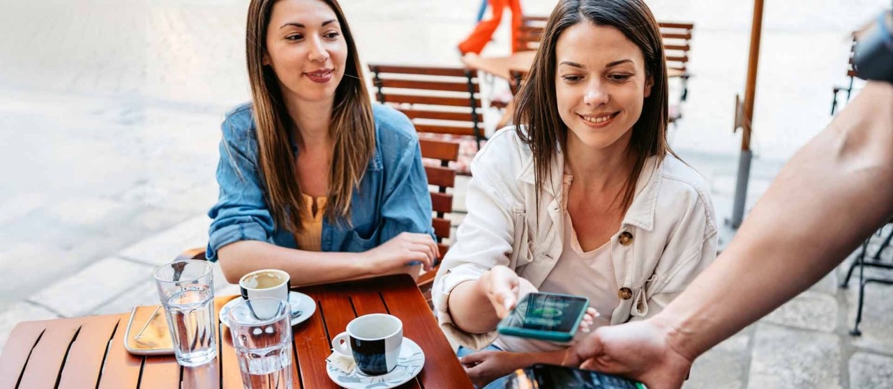 Dos personas en una cafetería al aire libre pagando con tarjeta mediante un dispositivo de cobro.