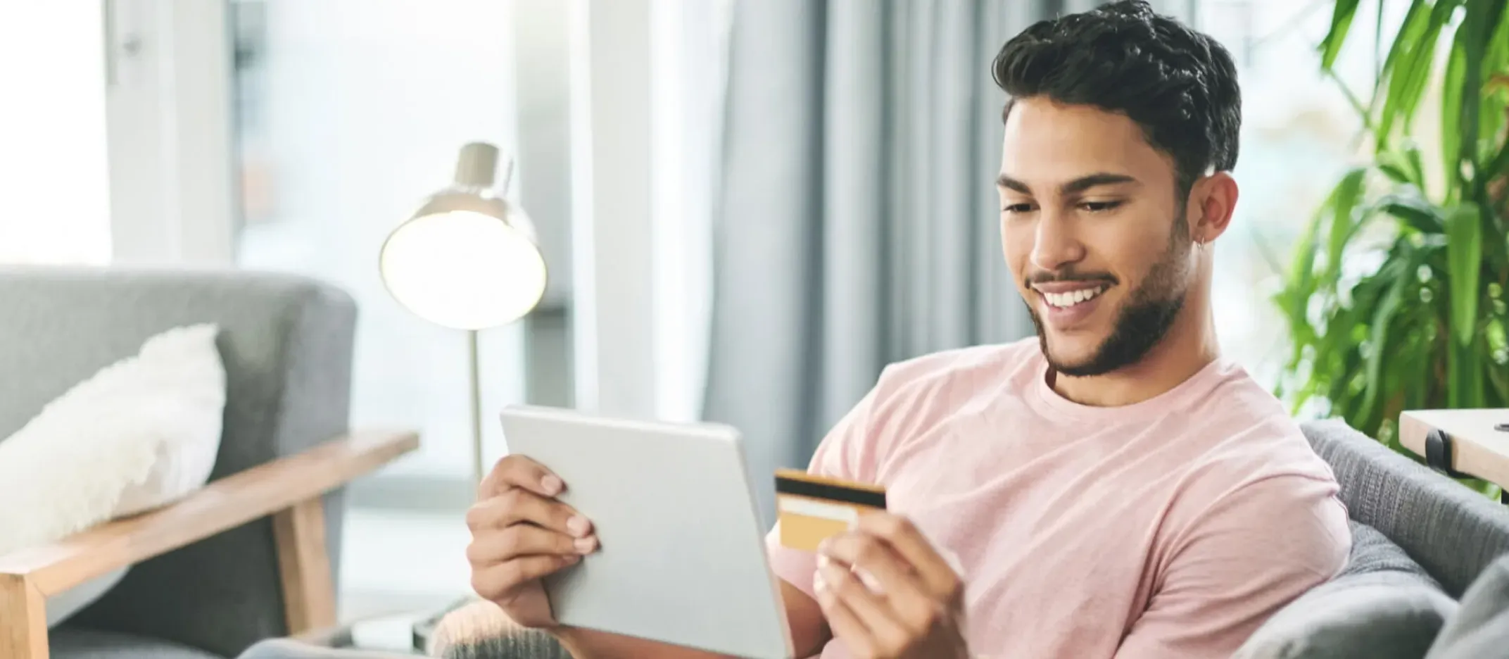 Persona sentada en sala sosteniendo tarjeta bancaria y tablet para realizar pago en línea.