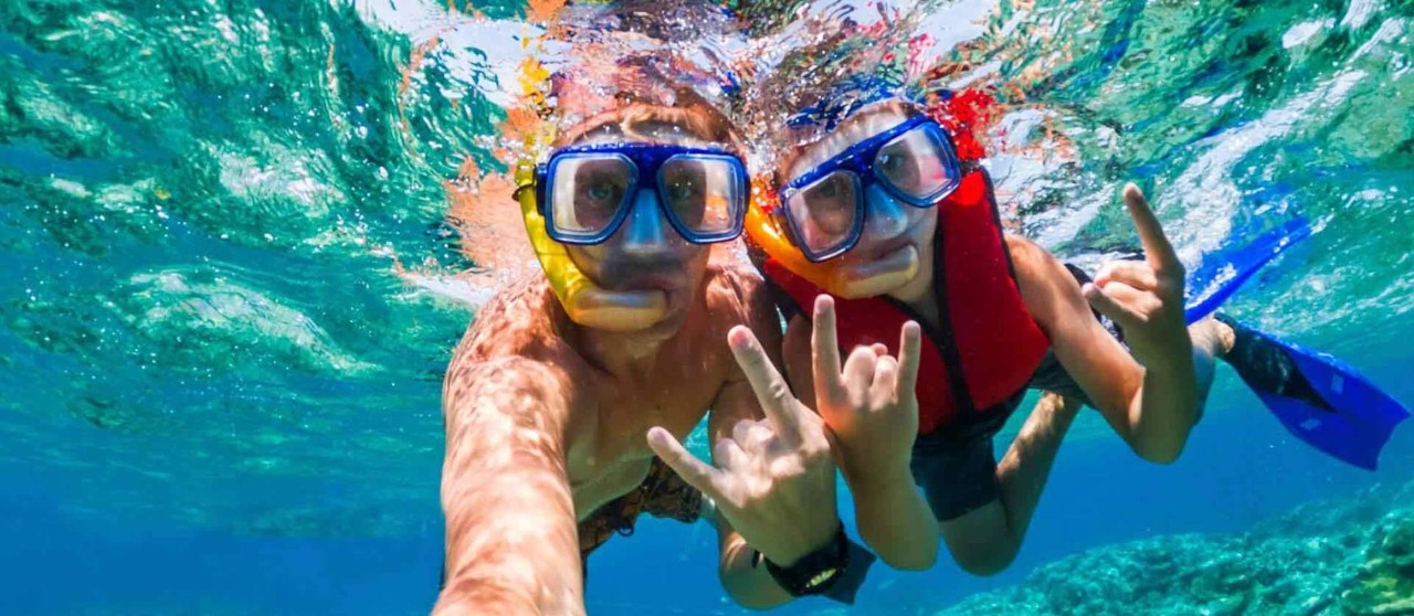 Dos personas practicando snorkel bajo el agua cristalina mostrando señas con las manos.