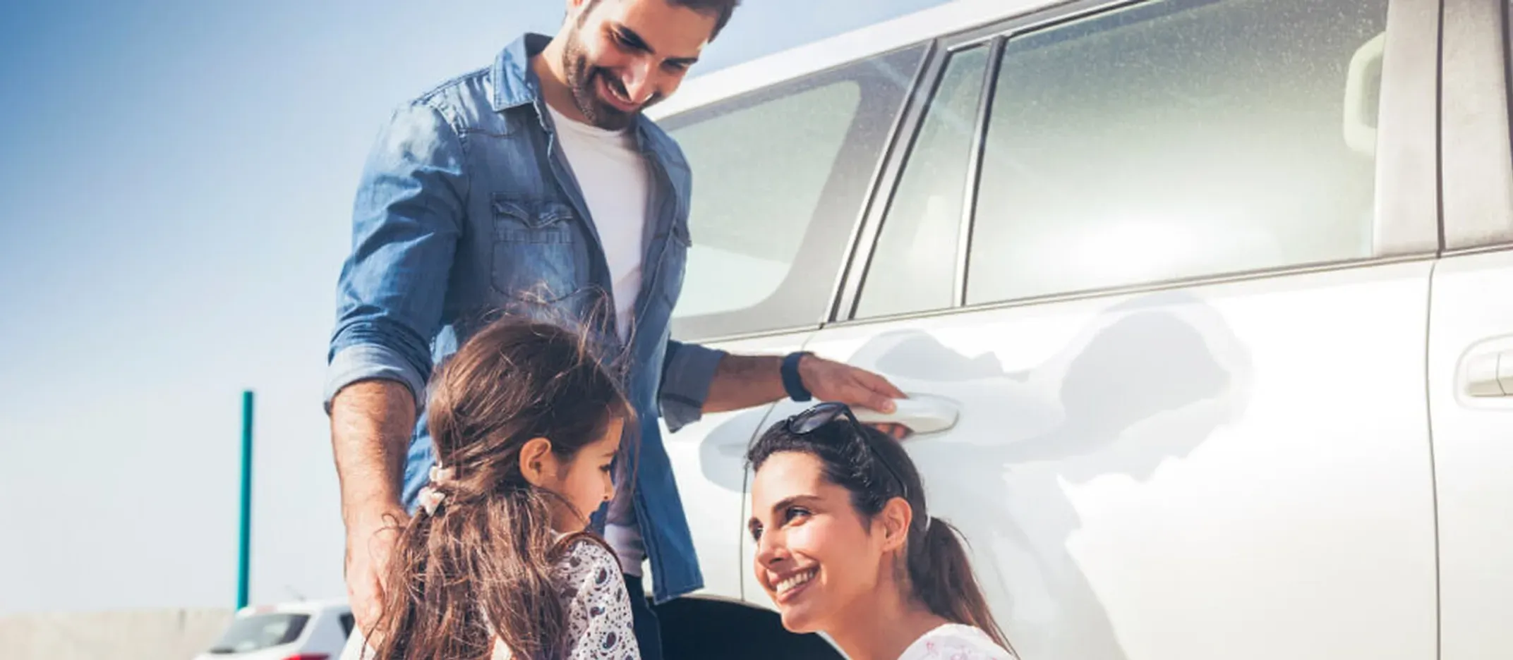 Familia preparando un viaje en carretera frente a un automóvil blanco en exterior soleado.