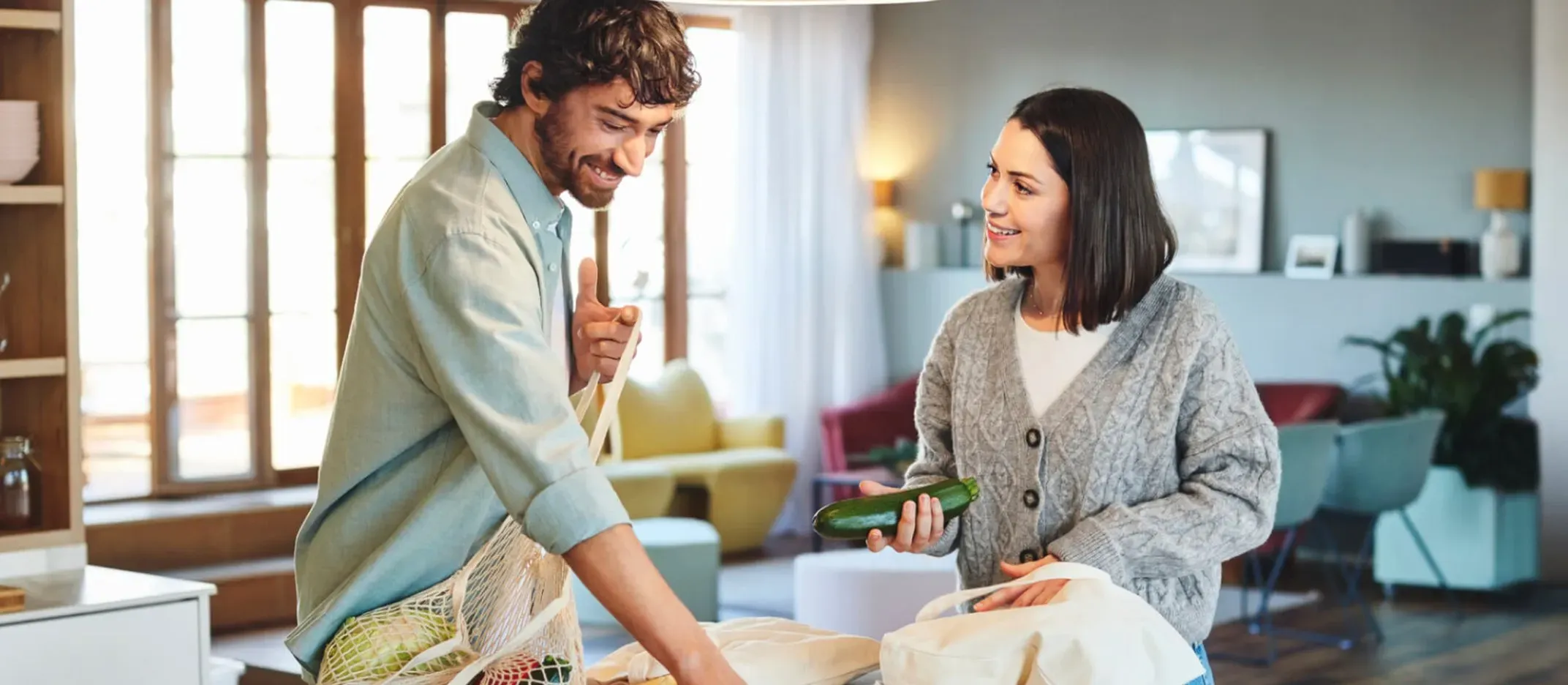 Dos personas colocando verduras sobre una mesa en una cocina iluminada por luz natural.