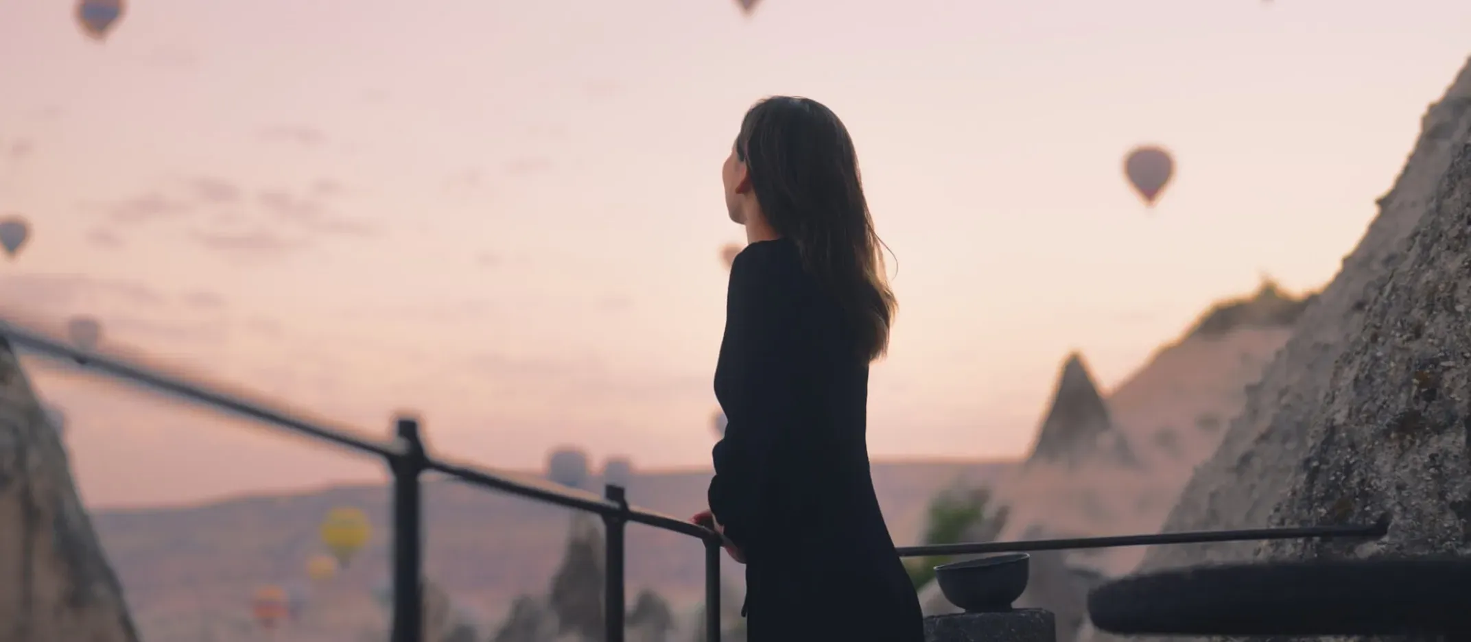 Persona contemplando globos aerostáticos al amanecer en un paisaje montañoso.