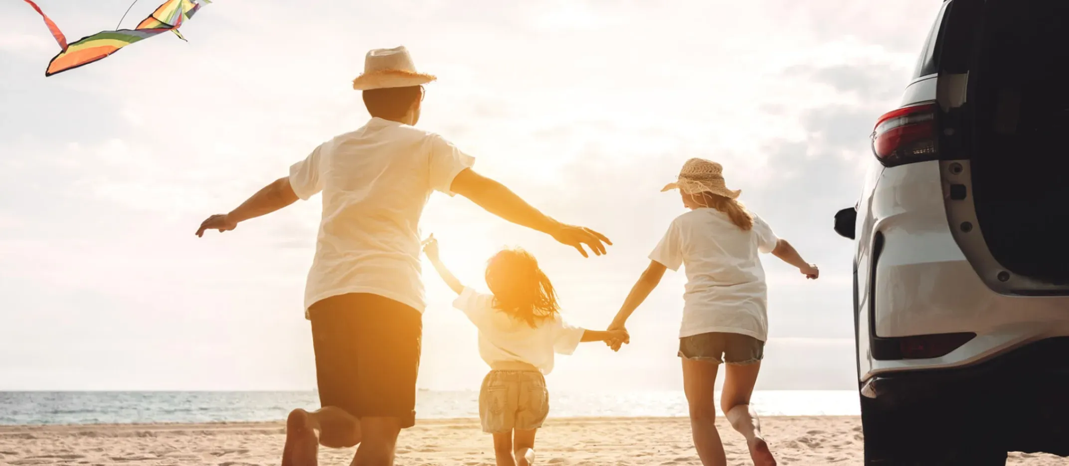 Familia corriendo en la playa junto a un auto mientras vuelan una cometa al atardecer.