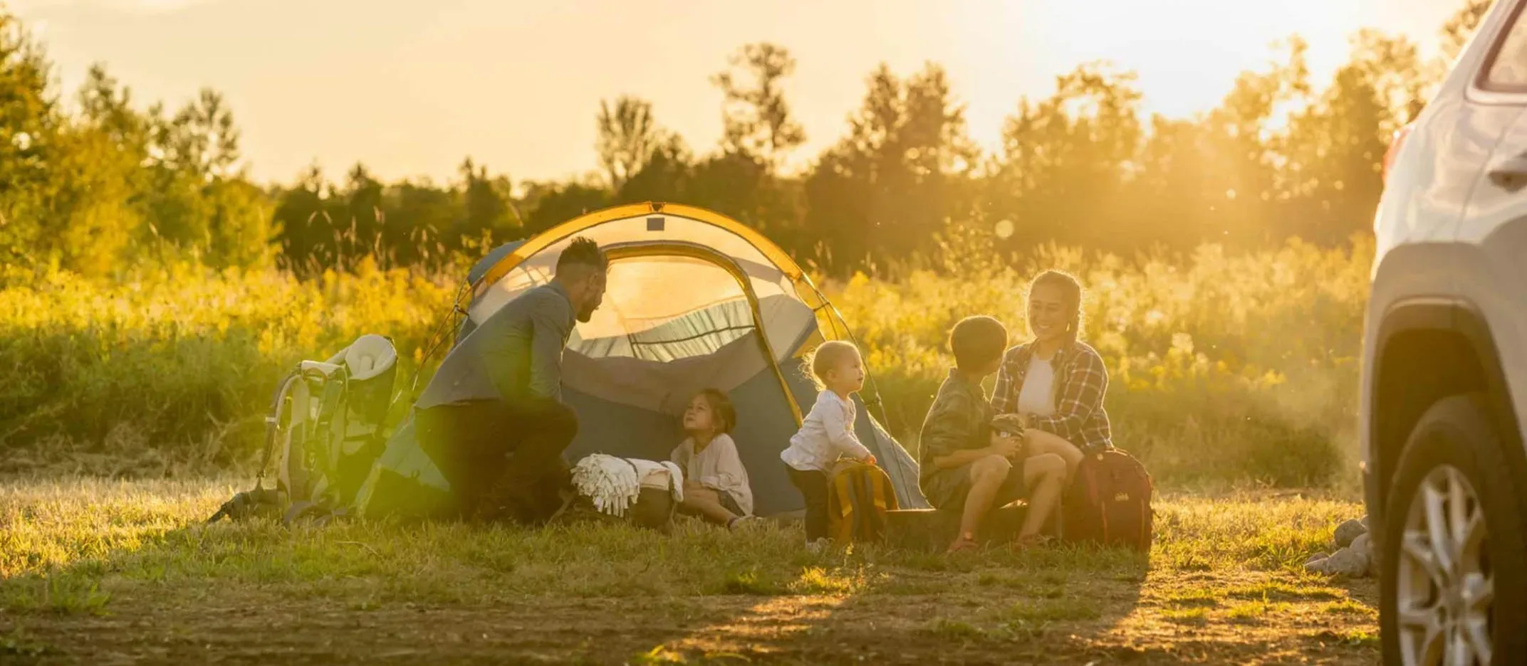 Familia acampando al aire libre junto a una tienda y un auto al atardecer.