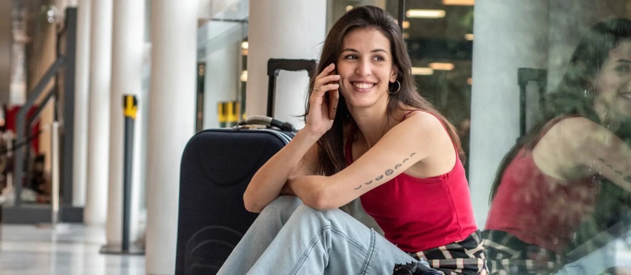 Persona sentada en el suelo de un aeropuerto junto a una maleta, hablando por teléfono cerca de una pared de vidrio.