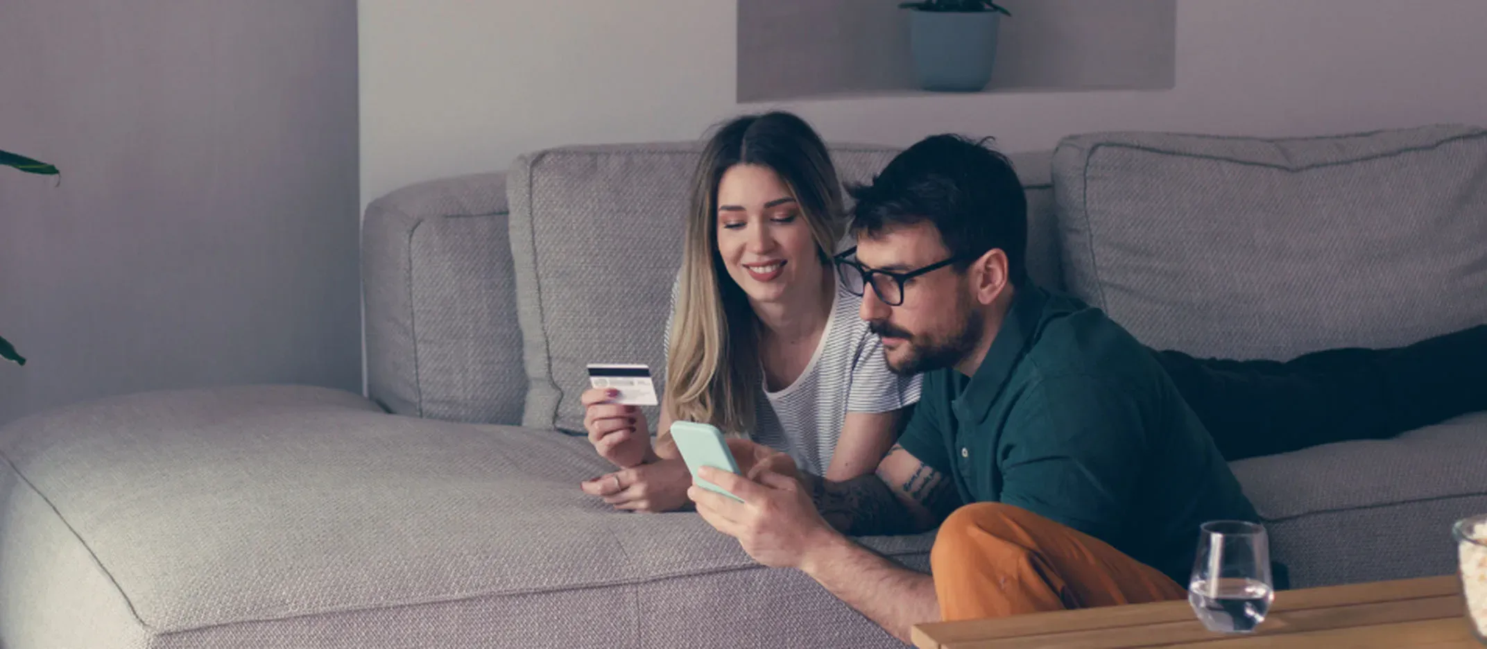 Dos personas en un sofá usando un teléfono móvil y una tarjeta bancaria para compras en línea.