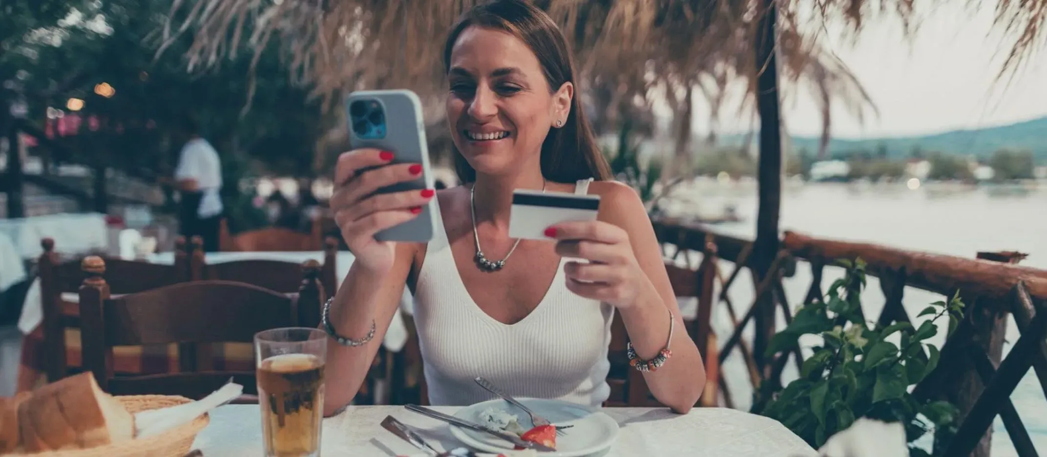 Persona en restaurante al aire libre sosteniendo un celular y una tarjeta de crédito sobre la mesa.