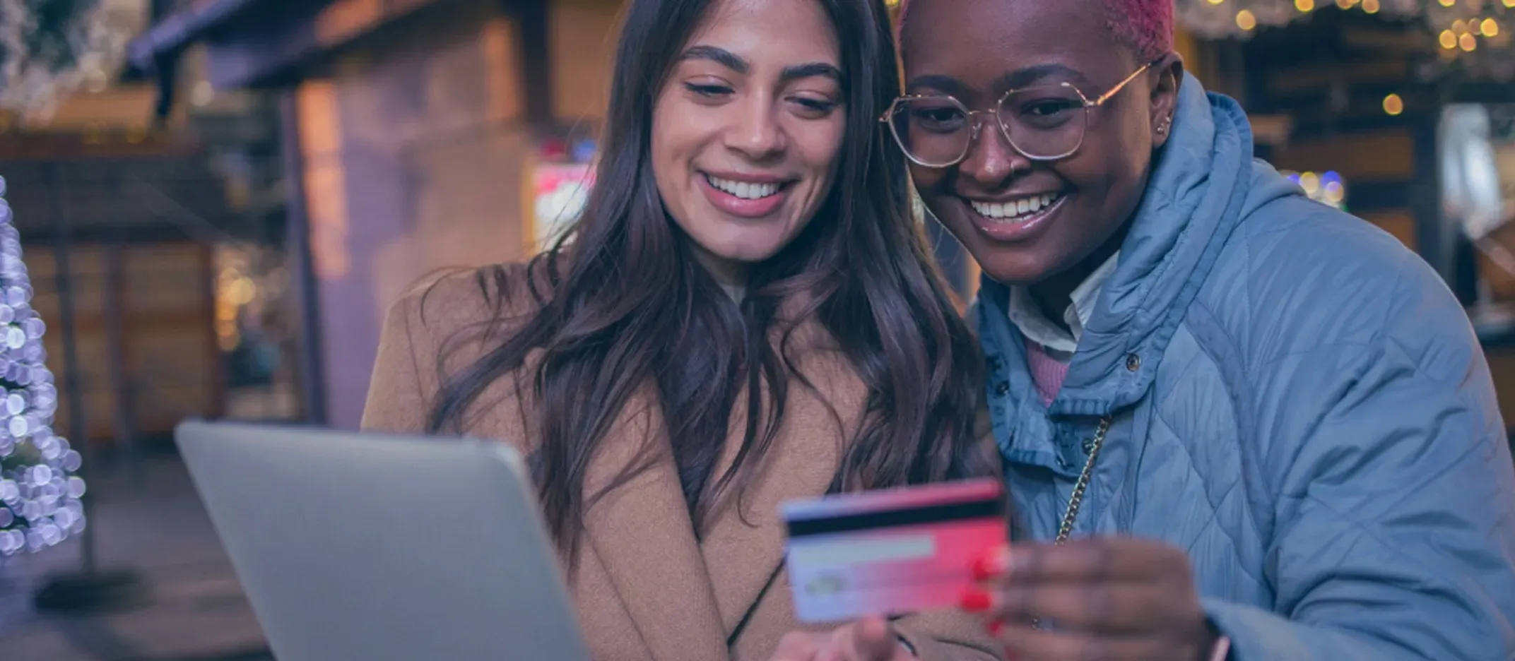 Dos personas al aire libre sosteniendo una tarjeta bancaria y usando una laptop con luces navideñas.