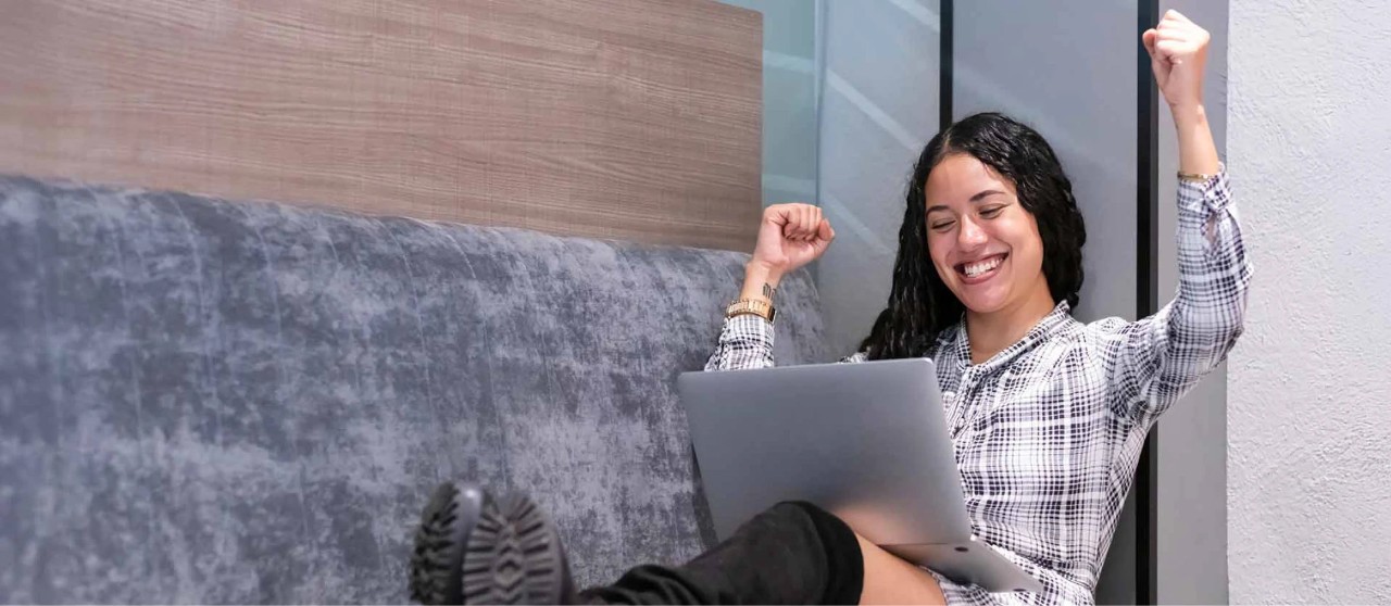 Persona sentada con una laptop levantando los brazos en señal de logro en un espacio interior moderno.