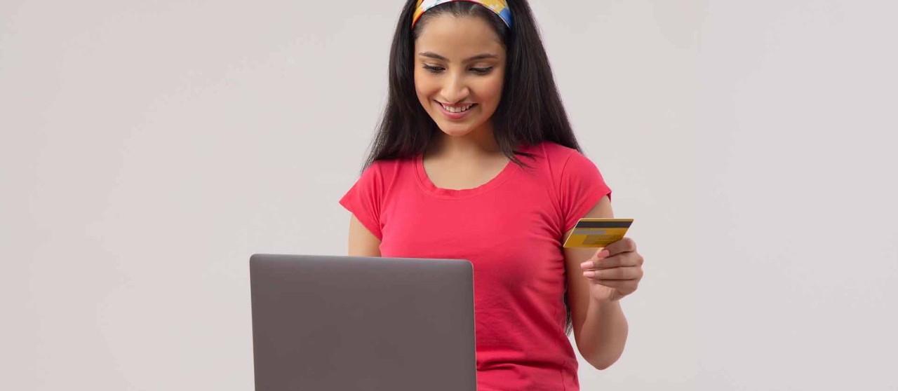 Persona usando una laptop y sosteniendo una tarjeta de crédito sobre fondo claro.