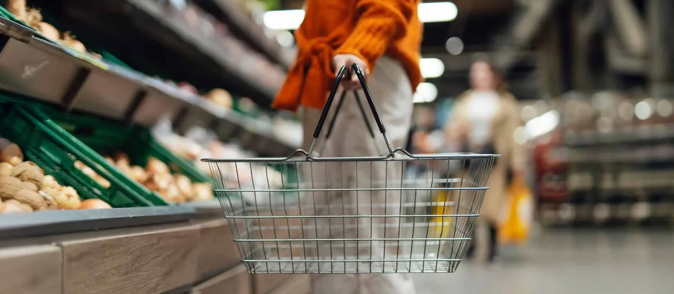 Persona sosteniendo una canasta de compras en el pasillo de frutas y verduras del supermercado.