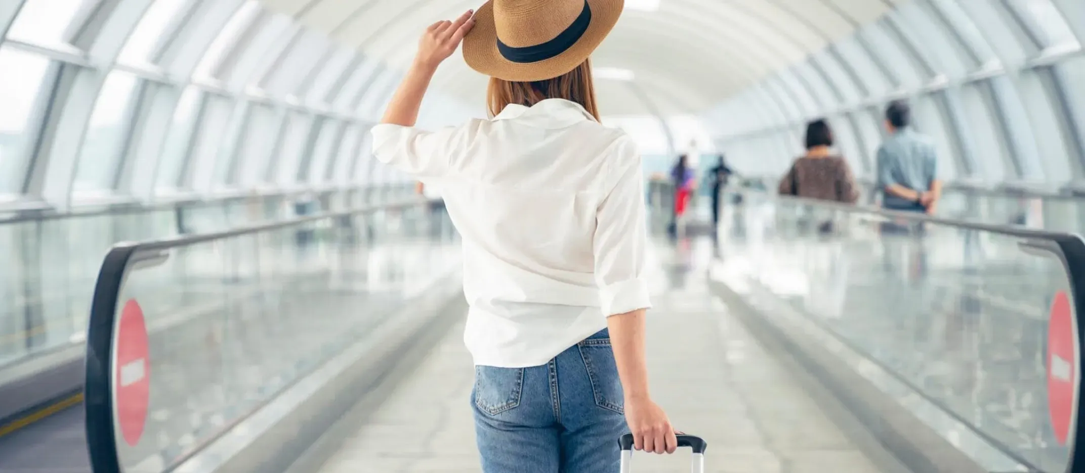 Persona caminando en un pasillo de aeropuerto con sombrero y maleta de mano.