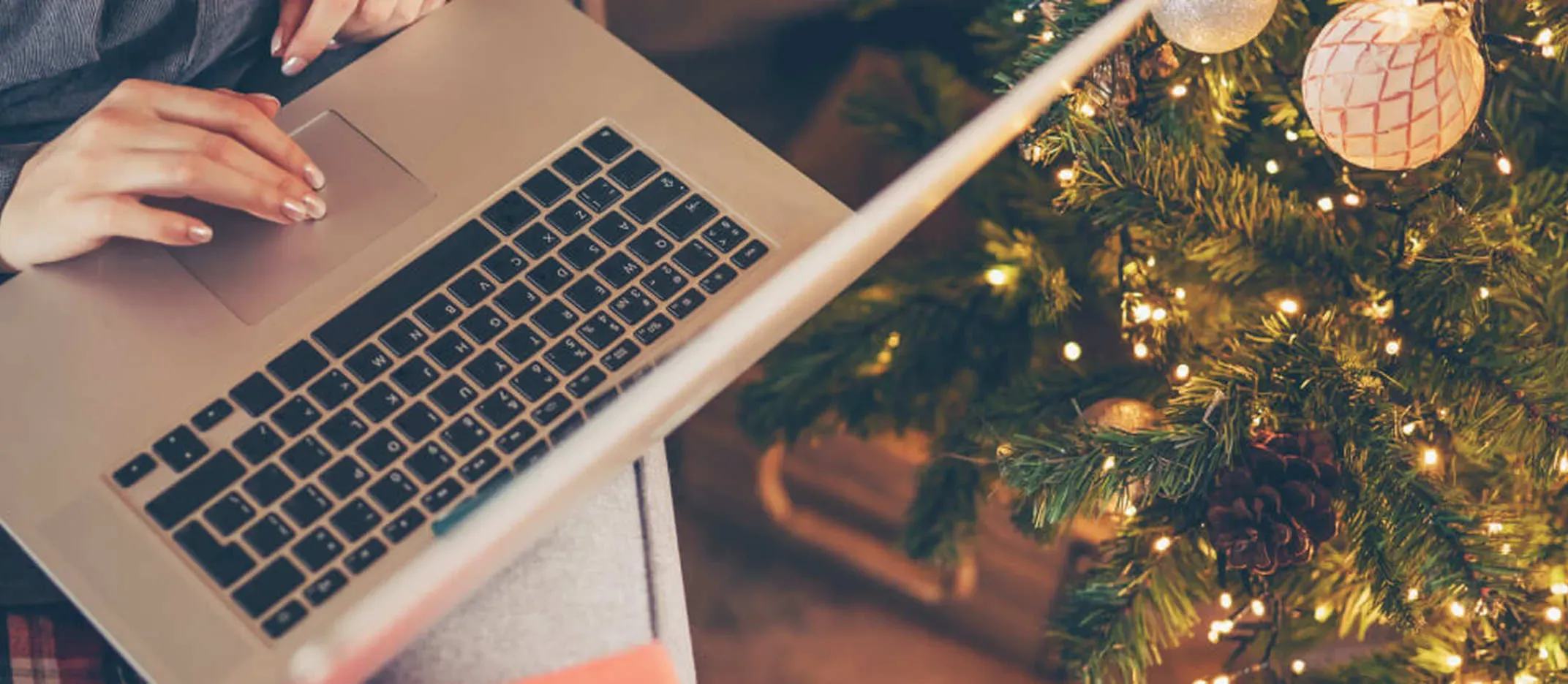 Persona usando laptop junto a árbol de Navidad decorado con luces y esferas.