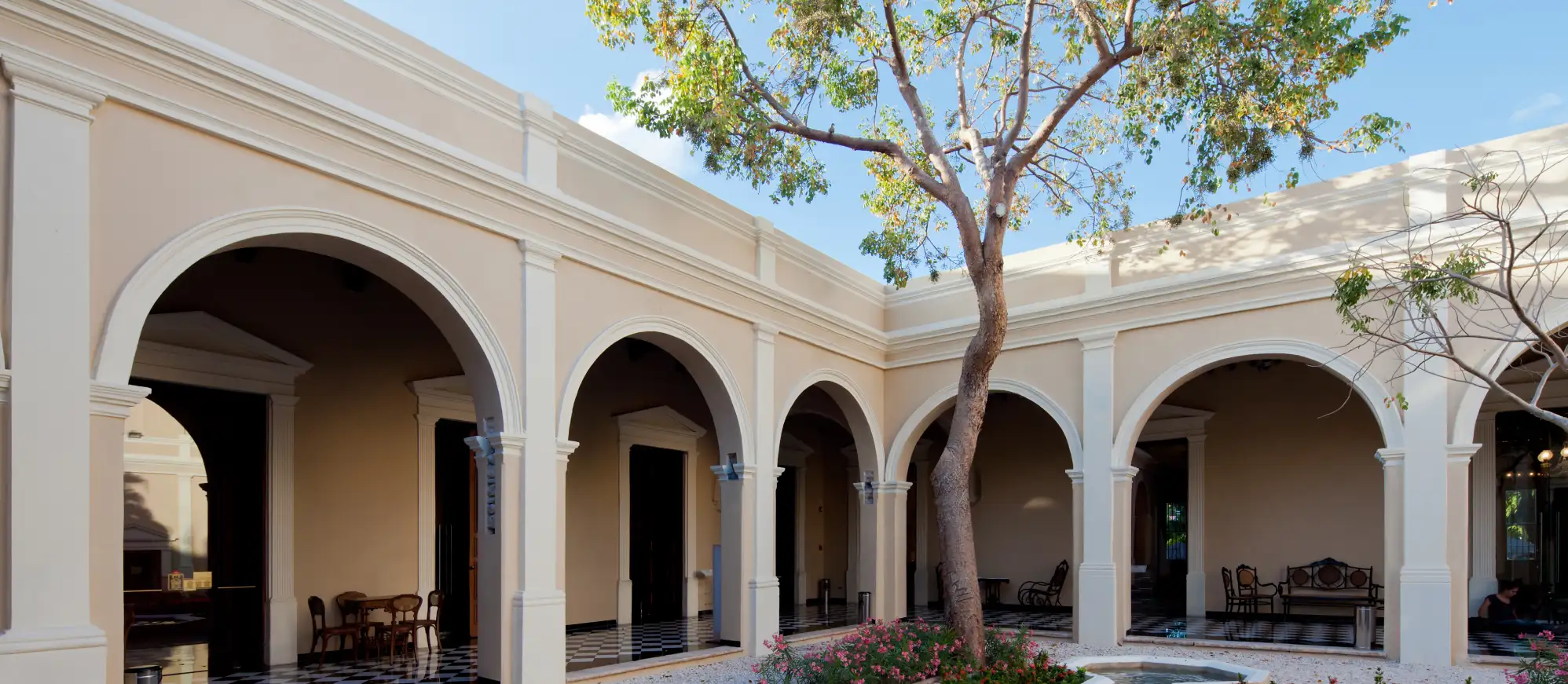 Patio con arcos, columnas y un árbol de flores rosas al centro en Casa Montejo, sede cultural de Banamex en Mérida