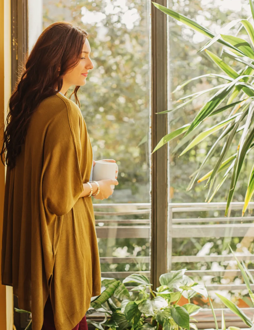Persona sosteniendo una taza junto a una ventana rodeada de plantas, representando tranquilidad, bienestar y la confianza de contar con soluciones financieras para el día a día con Banamex.