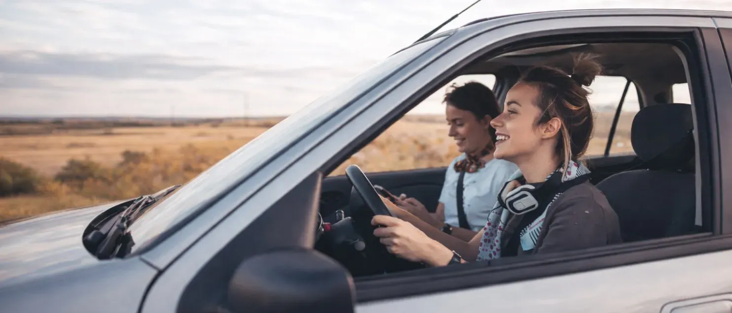 Una mujer conduciendo un automóvil mientras otra usa un celular en el asiento delantero.