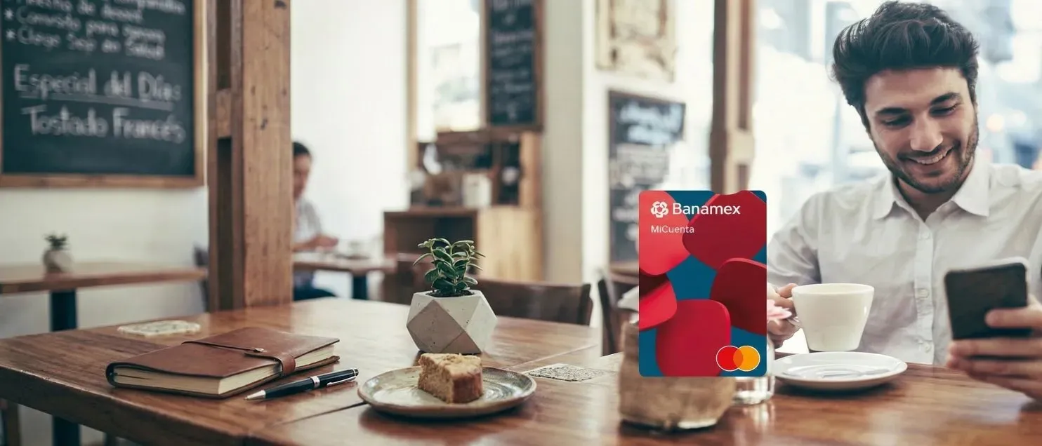 Persona sentada en una mesa de cafetería sosteniendo un teléfono móvil y una taza blanca, con una tarjeta Banamex MiCuenta Mastercard en primer plano.