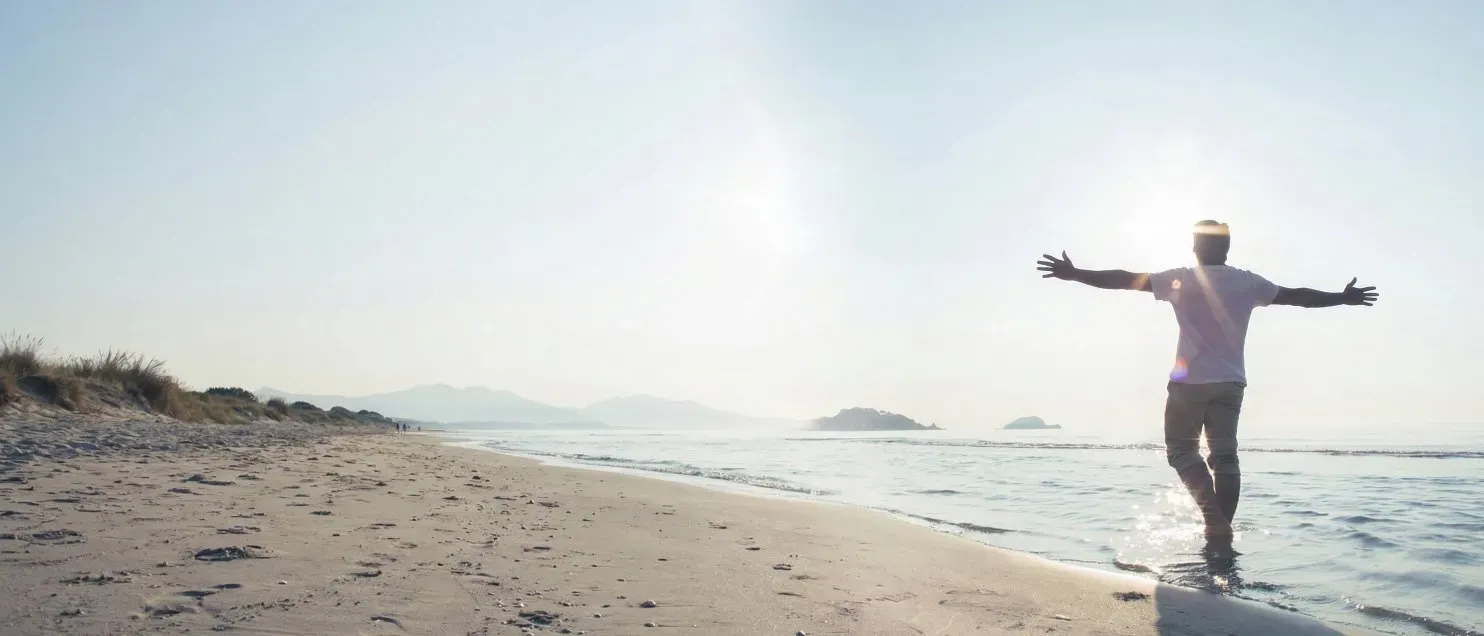 Persona con brazos abiertos en la orilla del mar al amanecer, simbolizando libertad financiera y tranquilidad para el retiro..