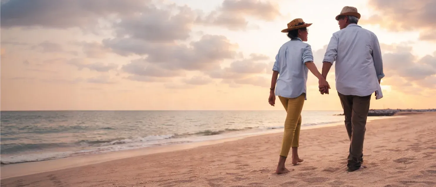 Pareja caminando de la mano por la playa al atardecer, simbolizando tranquilidad y bienestar financiero en la jubilación.