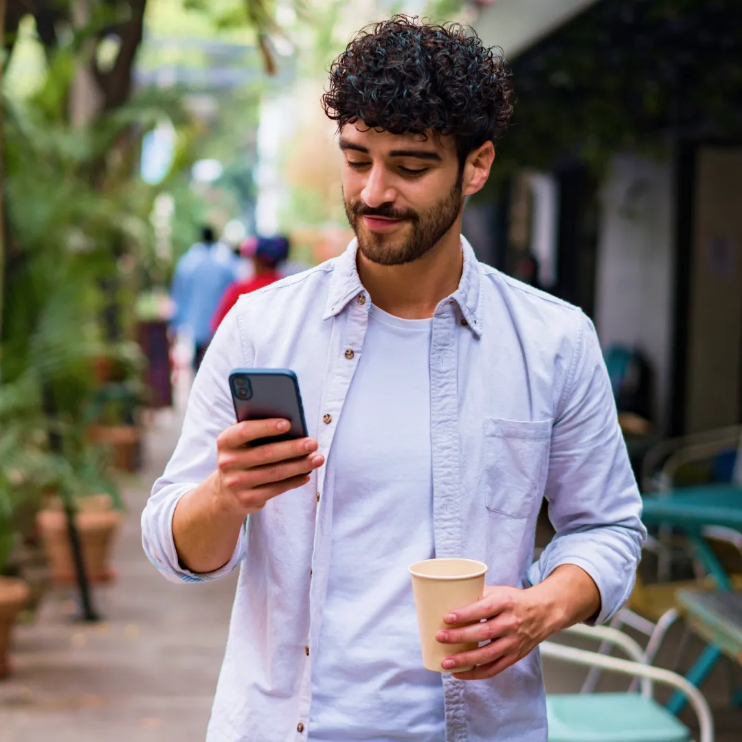 Persona caminando al aire libre mientras consulta un teléfono móvil y sostiene una bebida, representando el acceso sencillo a servicios financieros y banca digital Banamex desde cualquier lugar.