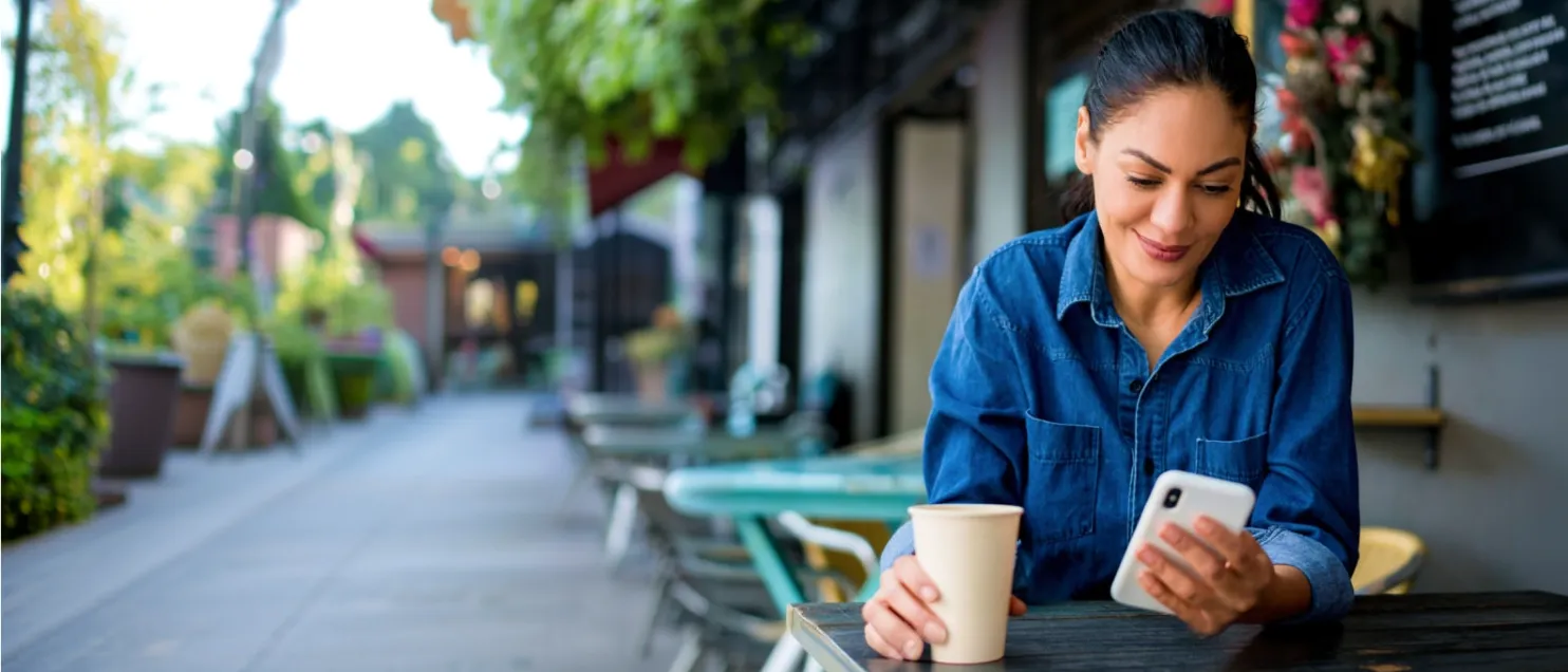 Persona utilizando un teléfono móvil mientras disfruta una bebida en una cafetería al aire libre, representando el acceso sencillo y seguro a servicios financieros digitales de Banamex.