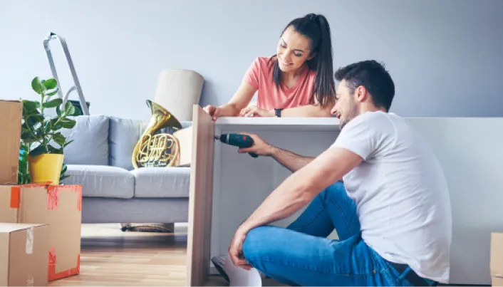 Pareja joven armando un mueble con un taladro entre cajas de mudanza en su nueva casa, representando la ilusión de estrenar hogar y el respaldo financiero de Banamex.