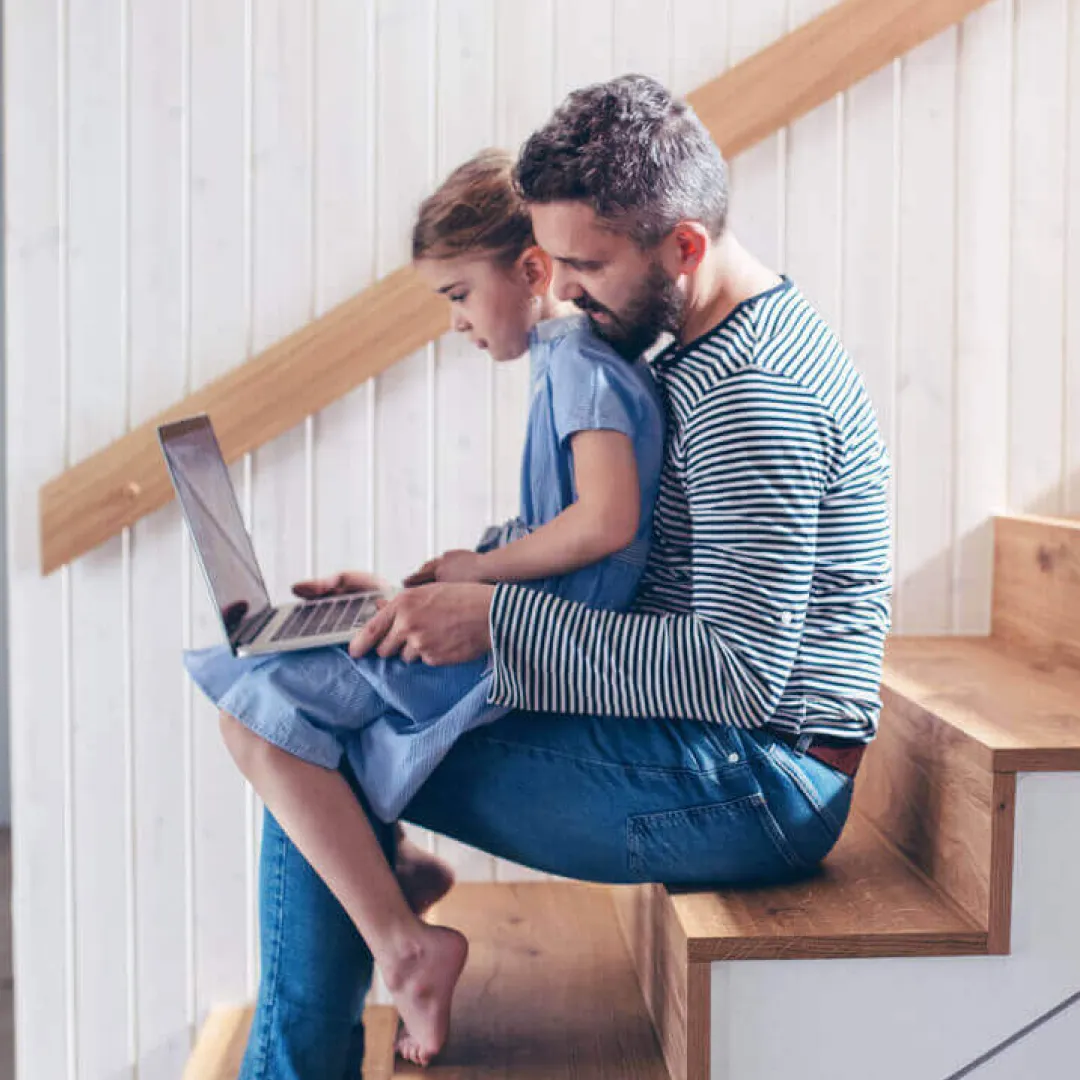 Padre e hija sentados en las escaleras de casa mirando juntos una laptop, representando la educación financiera en familia y el respaldo digital de Banamex.
