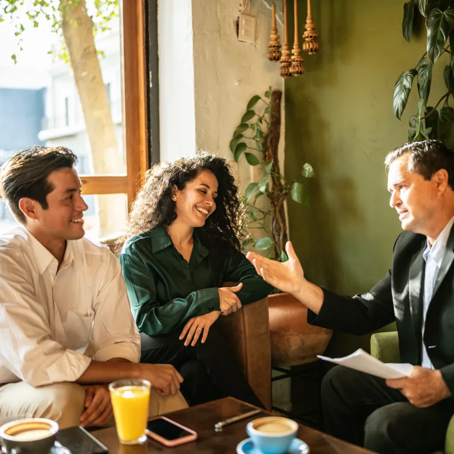 Asesor de Banamex conversando con una pareja sonriente en una cafetería, brindando asesoría financiera personalizada sobre créditos, inversiones o productos del banco.